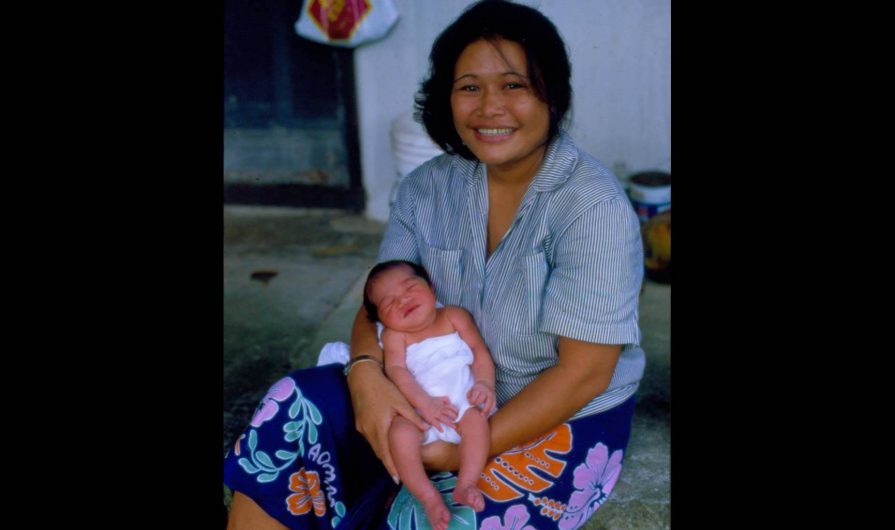 A Polynesian lady with a big smile, dressed in a bright sarong, holding a newborn baby.