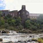 To the left is a large silver warehouse. Centre and right are large brick building covered in moss. There are a range of mostly dark green trees in front. At the very foreground is a river with large rocks.