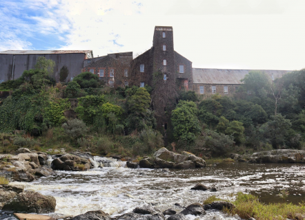 To the left is a large silver warehouse. Centre and right are large brick building covered in moss. There are a range of mostly dark green trees in front. At the very foreground is a river with large rocks.