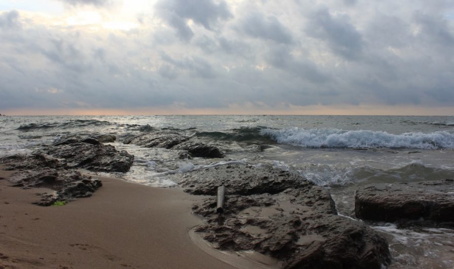 A beach with large rocks. The sand is wet and dark. There are small waves coming in and the sky is cloudy