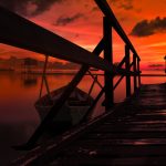 A wooden pier and small wooden boat looking over the ocean. The sunset is red and colouring everything in warm red colours.