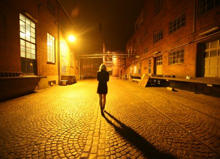 Woman in dark dress and medium light hair walking down street at night, facing away from camera. The street is lined with brick buildings and a bright yellow streetlight lights the woman and cobblestone path