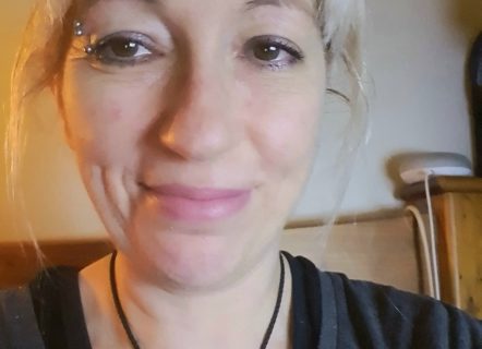 A woman with blonde hair pulled back into a ponytail and a silver eyebrow piercing. She is smiling and looking at the camera