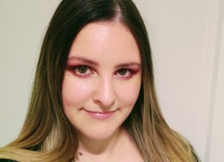A young woman with long dark hair that fades to blonde and a closed mouth smile. She is standing in front of a blank wall and wearing a black t-shirt