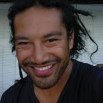 A close up photo of a man in a dark shirt with stubble and dreadlocks that are pulled back into a bun. He is smiling at the camera