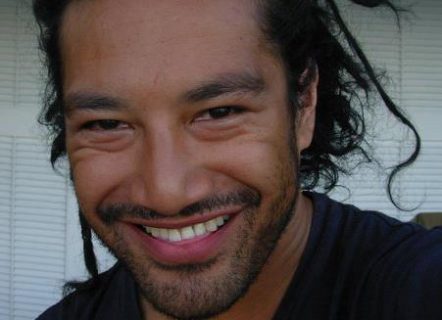 A close up photo of a man in a dark shirt with stubble and dreadlocks that are pulled back into a bun. He is smiling at the camera