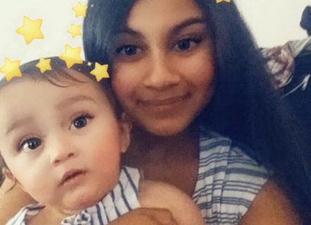 A young woman with dark long black hair is holding a small child with chubby cheeks and smiling at the camera. Both have a crown of small yellow stars across their heads