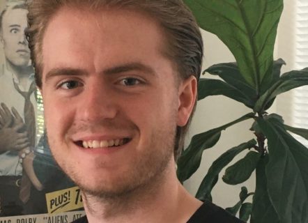 A man with a white face and pushed back dirty blonde hair stands smiling. He is in front of a large green plant and a poster of Thomas Dolby’s album cover Aliens ate my Buick, the man and woman look scared as an alien snake is emerging from a UFO eating a Buick