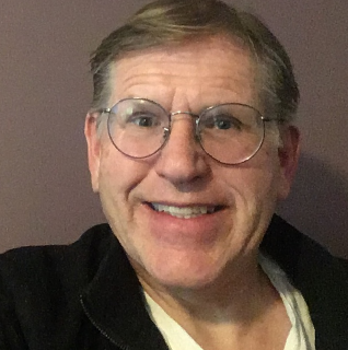 Self-portrait picture of a mature man. The picture is cut off at the chest and is taken before a grey background. He is wearing glasses. His hair is light blonde, slightly grey and is parted on the left side. The man is smiling with teeth. He is wearing glass, a black top with an open zip over a white t-shirt