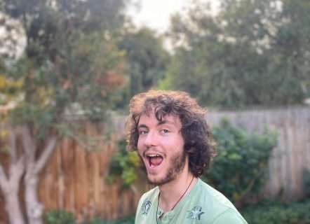 A youthful man with wild brown curls and a short scruffy beard. He is wearing a mint panthers t-shirt, smiling with his mouth wide open, and standing in an open backyard