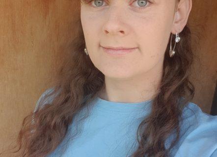 A woman wearing a blue shirt is standing in front of a brown backdrop. Her hair is long, light, curly and she has a fringe. She is looking at the camera and smiling