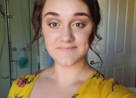A girl with curled brown hair, full brown eyebrows and hazel eyes, wears a yellow dress with a pink flower pattern on it. Her minimal eye makeup matches the yellow of her dress. She stands in front of a window with a shower behind her, taking a photo looking into the camera lens. The dimple on her cheek is showing as she smiles for the photo