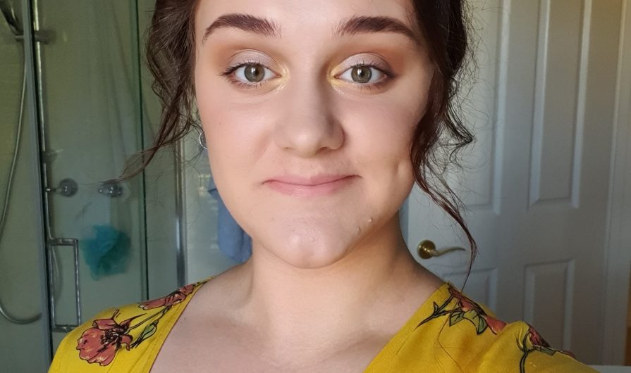 A girl with curled brown hair, full brown eyebrows and hazel eyes, wears a yellow dress with a pink flower pattern on it. Her minimal eye makeup matches the yellow of her dress. She stands in front of a window with a shower behind her, taking a photo looking into the camera lens. The dimple on her cheek is showing as she smiles for the photo