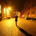 Woman in dark dress and medium light hair walking down street at night, facing away from camera. The street is lined with brick buildings and a bright yellow streetlight lights the woman and cobblestone path